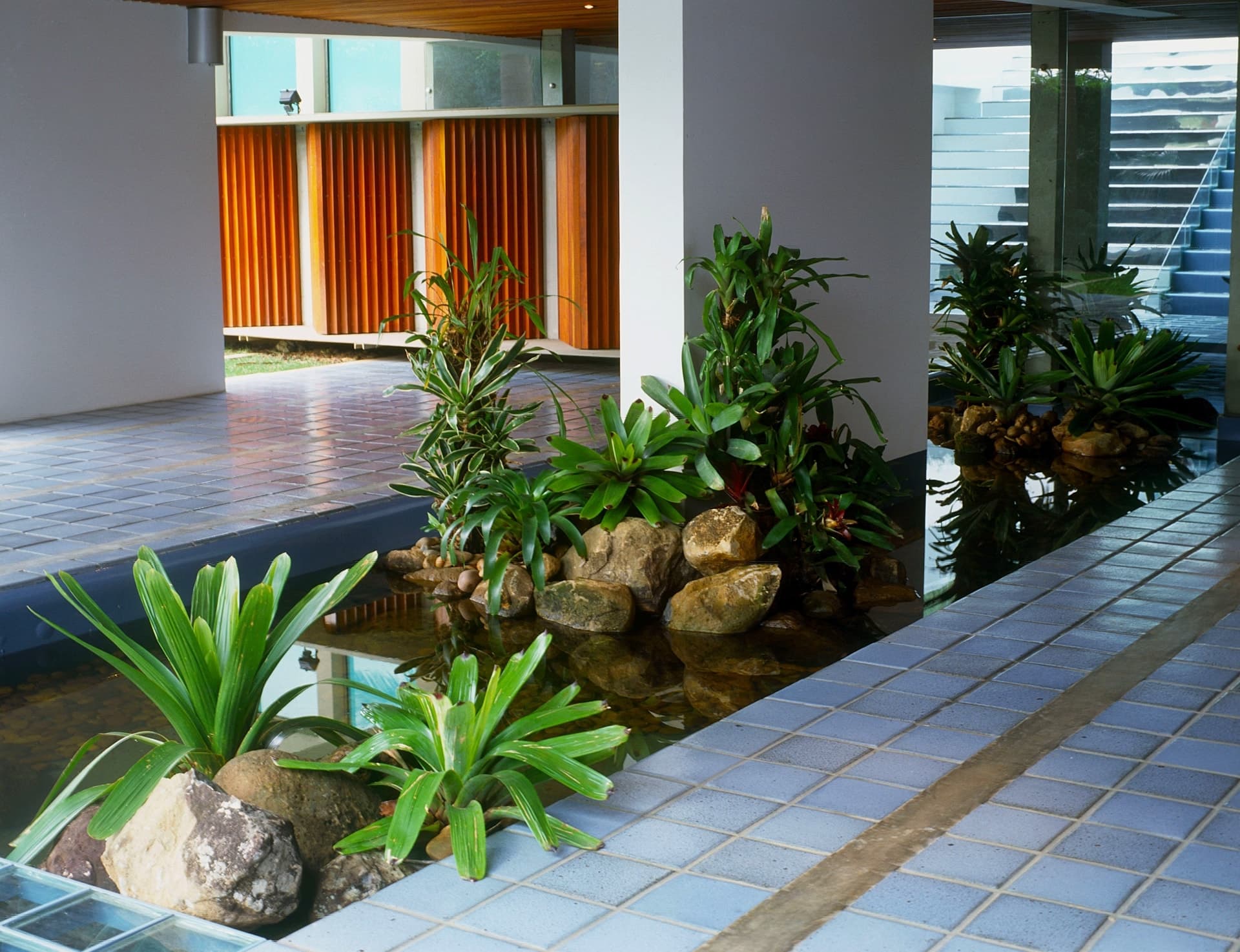 Jardim tropical com bromélias, pedras roladas e espelho d’água